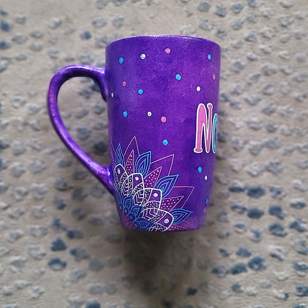 A Purple Hand painted mug with corner mandalas drawing on each of the mug corners in white pinks and blues and the name Noura in the center