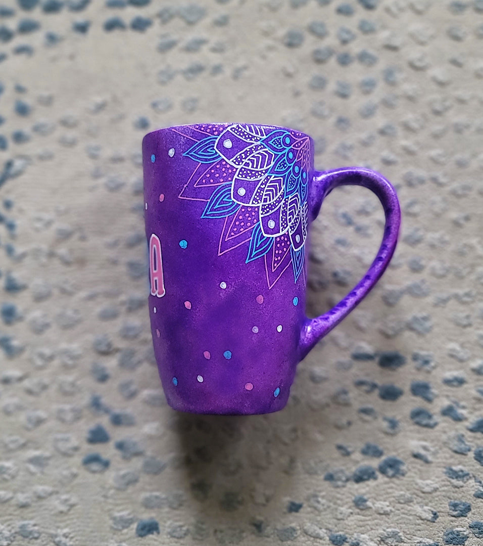 A Purple Hand painted mug with corner mandalas drawing on each of the mug corners in white pinks and blues and the name Noura in the center