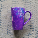 A Purple Hand painted mug with corner mandalas drawing on each of the mug corners in white pinks and blues and the name Noura in the center