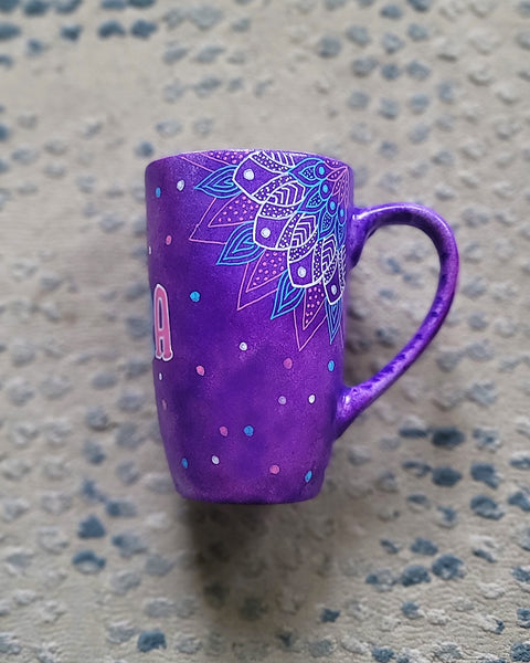 A Purple Hand painted mug with corner mandalas drawing on each of the mug corners in white pinks and blues and the name Noura in the center