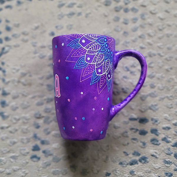 A Purple Hand painted mug with corner mandalas drawing on each of the mug corners in white pinks and blues and the name Noura in the center