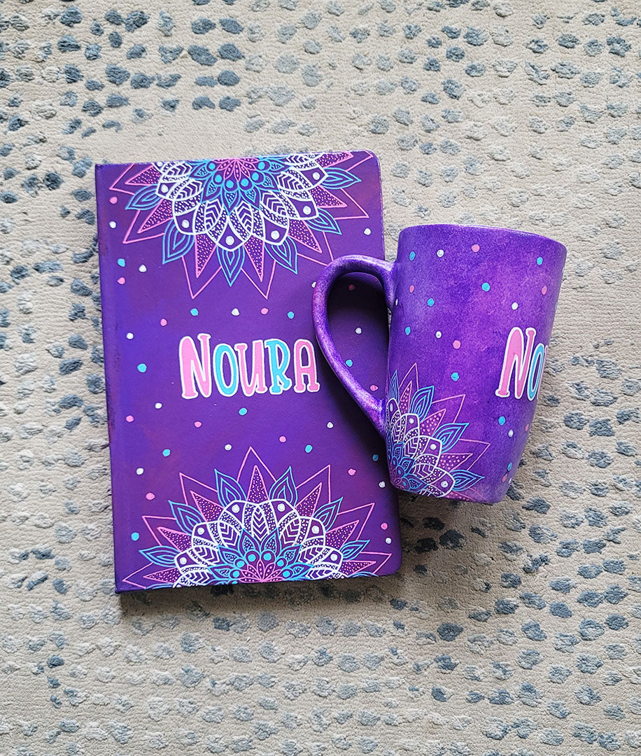 A Purple Hand painted gift set containing mug and notebook with the mug having corner mandalas and teh notebook having 1/2 mandalas on top and bottom and in between both the name "Noura"