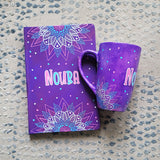 A Purple Hand painted gift set containing mug and notebook with the mug having corner mandalas and teh notebook having 1/2 mandalas on top and bottom and in between both the name 