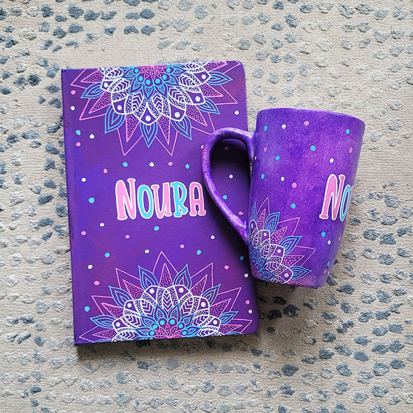 A Purple Hand painted gift set containing mug and notebook with the mug having corner mandalas and teh notebook having 1/2 mandalas on top and bottom and in between both the name "Noura"