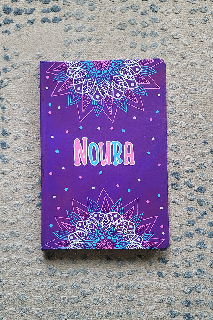 A Purple hand painted notebook with 2 half mandalas one on top and one at the bottom with the name "Noura" in the center