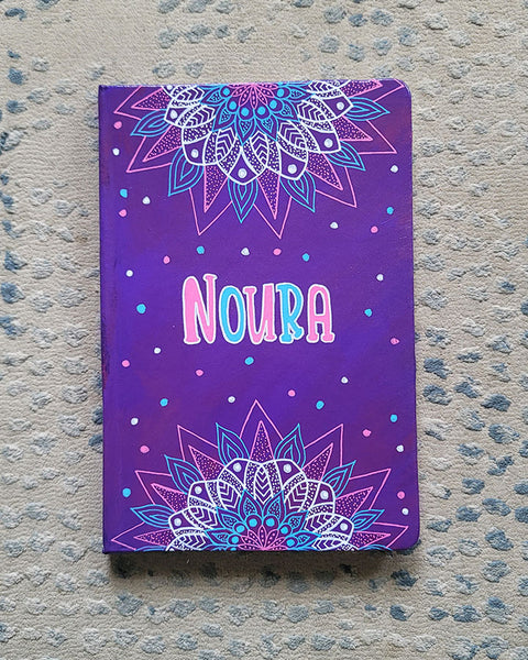 A Purple hand painted notebook with 2 half mandalas one on top and one at the bottom with the name "Noura" in the center