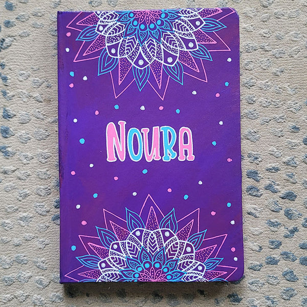 A Purple hand painted notebook with 2 half mandalas one on top and one at the bottom with the name "Noura" in the center