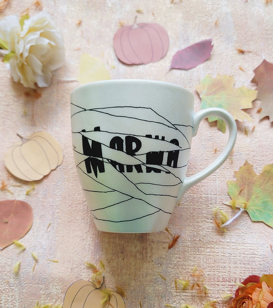 an ivory hand painted mug with a mummy drawn all over it and the name 'Marwa' peaking from behind