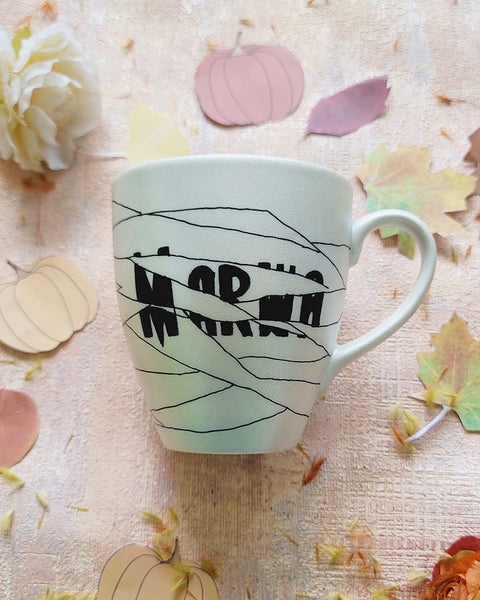 an ivory hand painted mug with a mummy drawn all over it and the name 'Marwa' peaking from behind