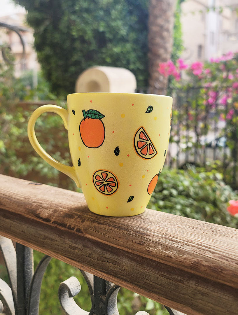 A Pale yellow hand painted mug with complete oranges and orange slices drawn in black and colored in with several degrees of orange color, with small green leaves and orange and yellow dots scattered all over