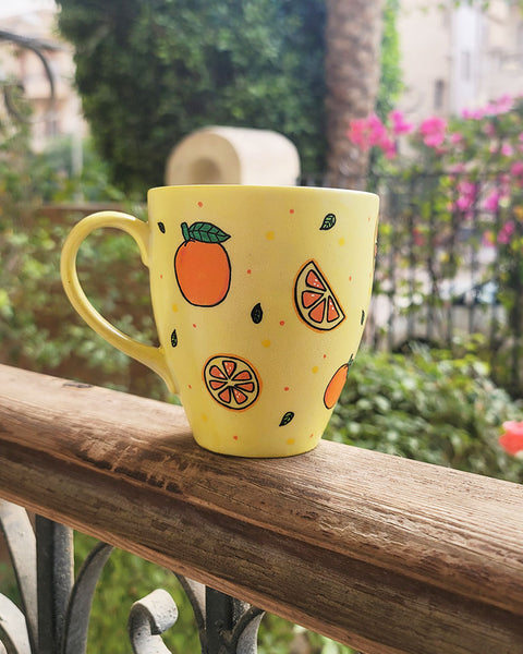 A Pale yellow hand painted mug with complete oranges and orange slices drawn in black and colored in with several degrees of orange color, with small green leaves and orange and yellow dots scattered all over