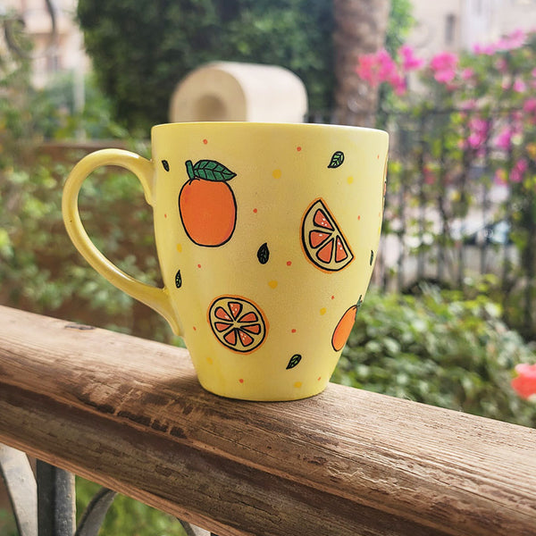 A Pale yellow hand painted mug with complete oranges and orange slices drawn in black and colored in with several degrees of orange color, with small green leaves and orange and yellow dots scattered all over