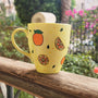 A Pale yellow hand painted mug with complete oranges and orange slices drawn in black and colored in with several degrees of orange color, with small green leaves and orange and yellow dots scattered all over