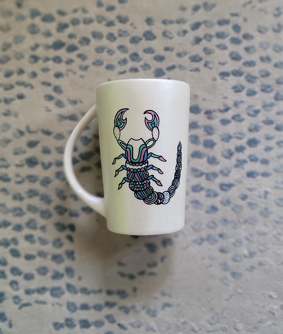 A Pearl white Hand painted Mug With a Scorpio Design on it in Black and colored in pinks, greens and blues