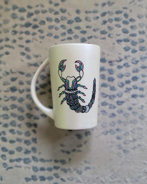 A Pearl white Hand painted Mug With a Scorpio Design on it in Black and colored in pinks, greens and blues