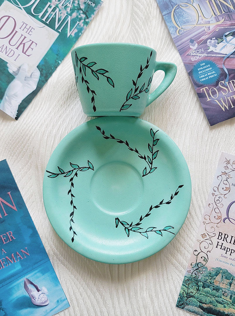 A Mint green hand painted cup and saucer with black relief leaves drawn across both the cup and plate and some leaves filled with shimmery mint.