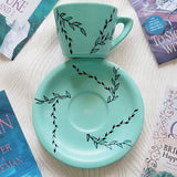 A Mint green hand painted cup and saucer with black relief leaves drawn across both the cup and plate and some leaves filled with shimmery mint.