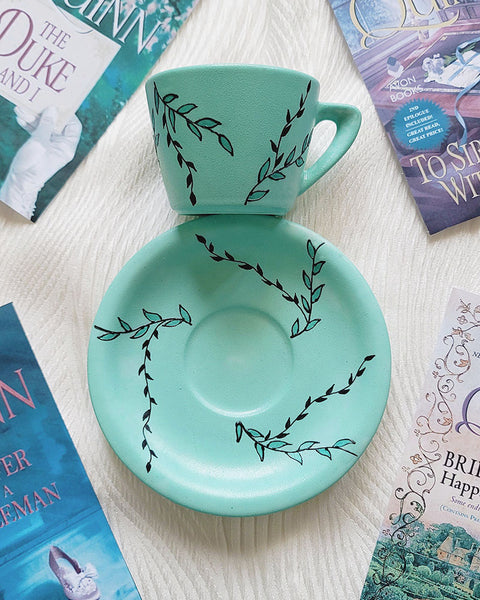 A Mint green hand painted cup and saucer with black relief leaves drawn across both the cup and plate and some leaves filled with shimmery mint.