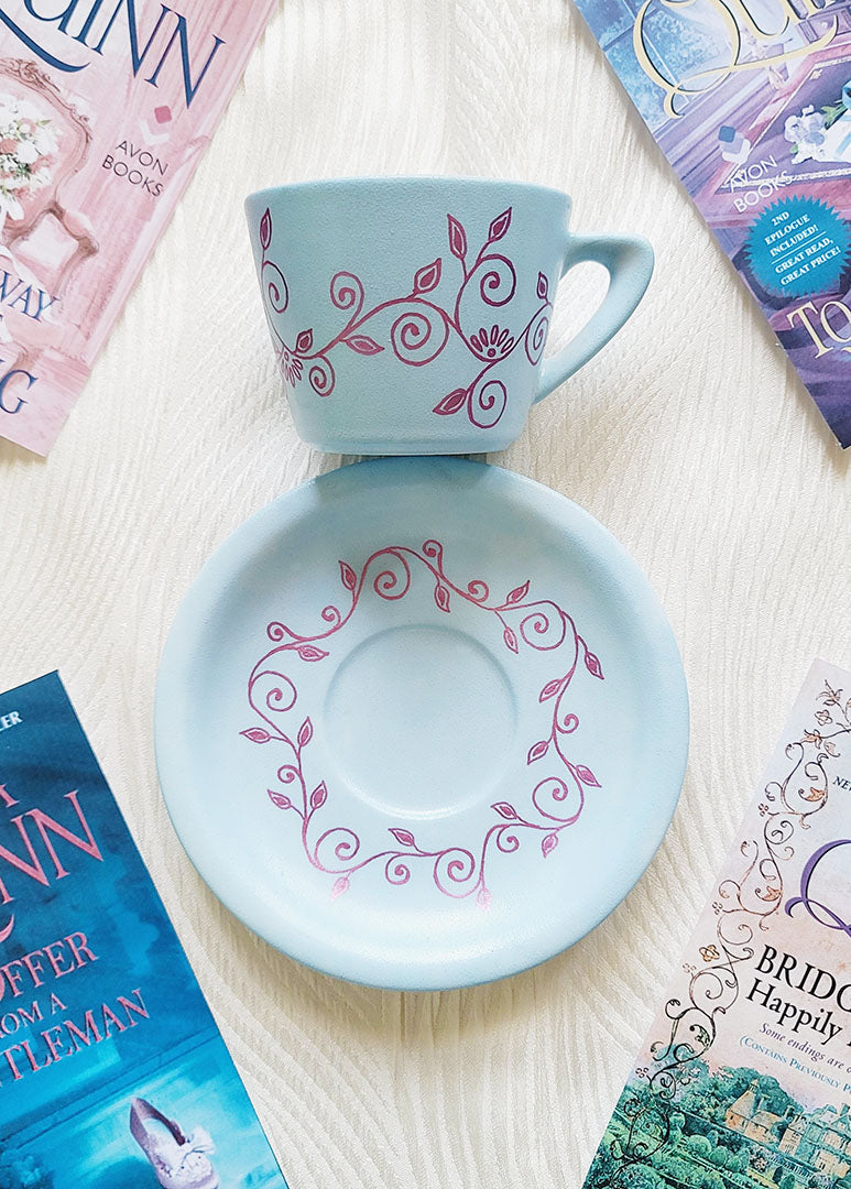 A Pale blue hand painted cup and saucer with lines of curves and leaves drawn all across the plate and cup in shimmery pink