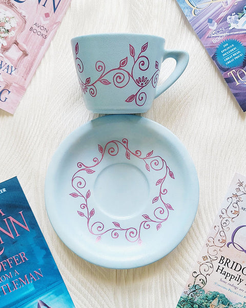 A Pale blue hand painted cup and saucer with lines of curves and leaves drawn all across the plate and cup in shimmery pink