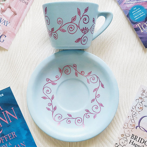 A Pale blue hand painted cup and saucer with lines of curves and leaves drawn all across the plate and cup in shimmery pink