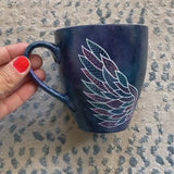 A black galaxy hand painted mug with a wing drawn on one side and the name 