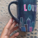 A Dark Sky handpainted mug with buildings drawn all over at the bottom half and the words 