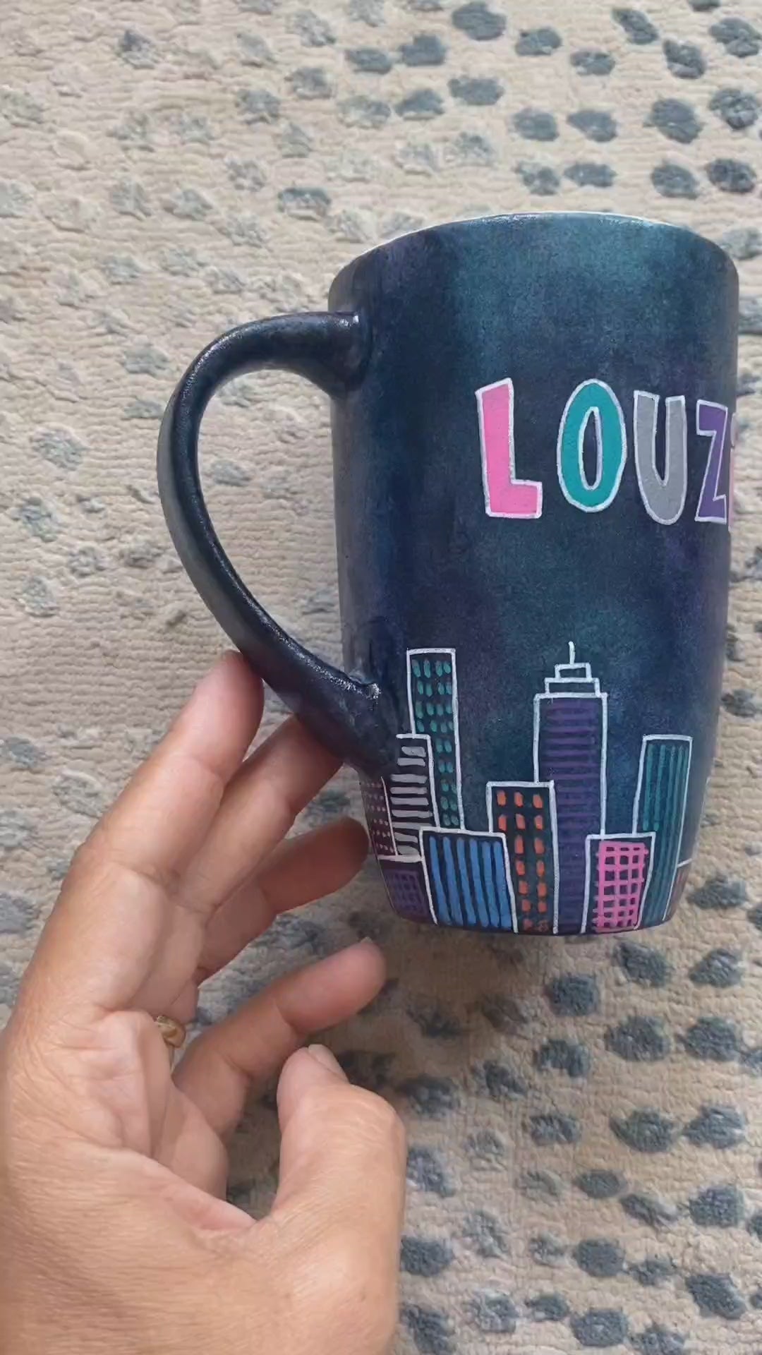 A Dark Sky handpainted mug with buildings drawn all over at the bottom half and the words "Finally and architect" and the other side has the name "Louzi"