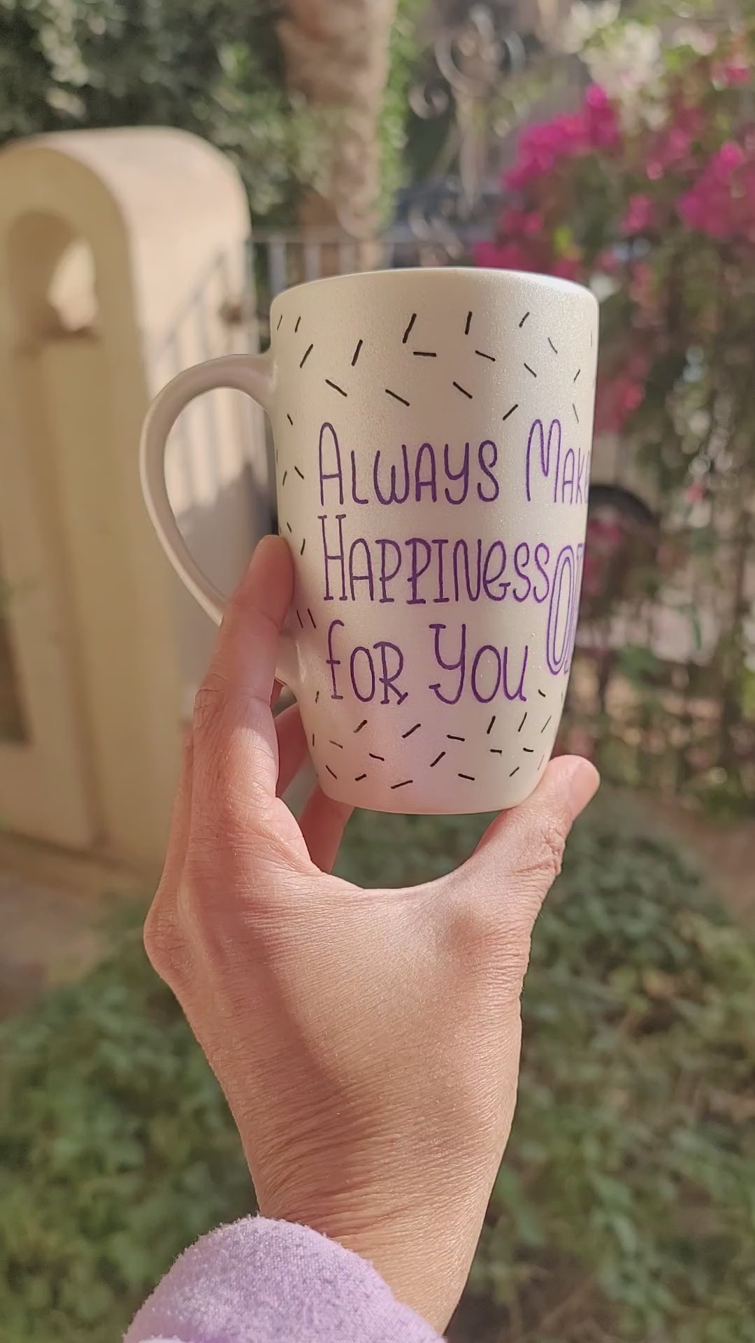 A white pearl hand painted mug with the words "Always Make Happiness ok for you " in purple and black sprinkles scattered all over the mug and the name "amira" on the other side