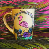 A Pastel yellow hand painted mug with a flamingo standing in the center over colorful flowers and around it half a mandala wreth. and on the other side the name 