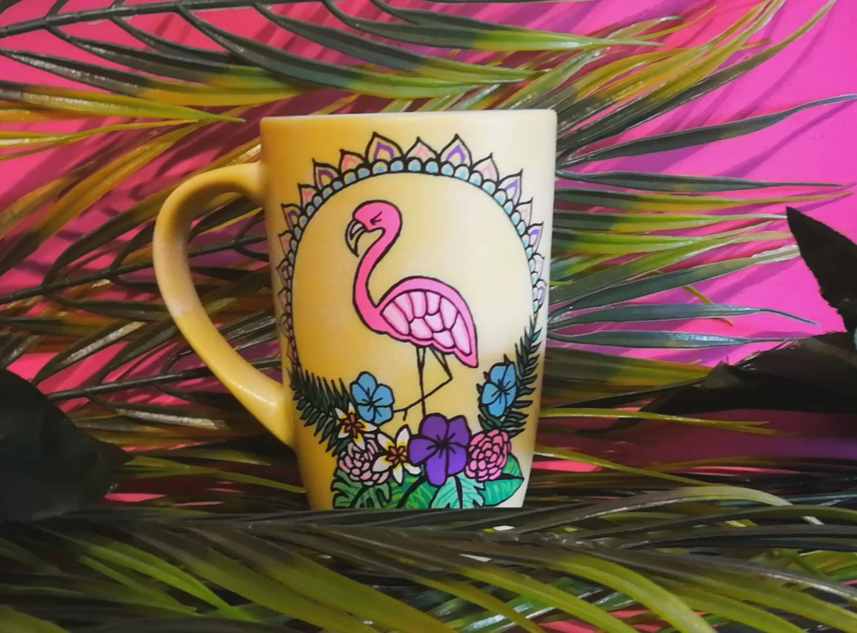 A Pastel yellow hand painted mug with a flamingo standing in the center over colorful flowers and around it half a mandala wreth. and on the other side the name "yara"