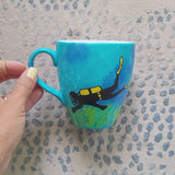 A Blue hand painted mug with a diver painted on it and corals all over the bottom half of the mug and the other side has the name 