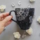 A bLack Galaxy hand painted mug with the name rousha in the middle and ont he corners a mandala drawn in white with patterns on dark grey and light grey. And light grey and dark grey dots scattered all over the mug