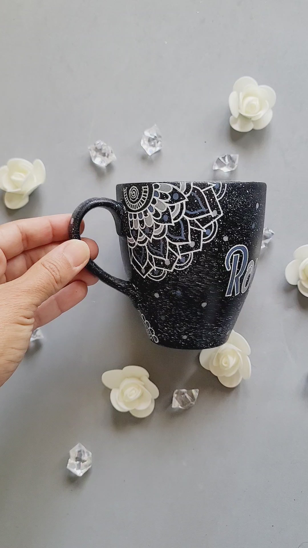 A bLack Galaxy hand painted mug with the name rousha in the middle and ont he corners a mandala drawn in white with patterns on dark grey and light grey. And light grey and dark grey dots scattered all over the mug