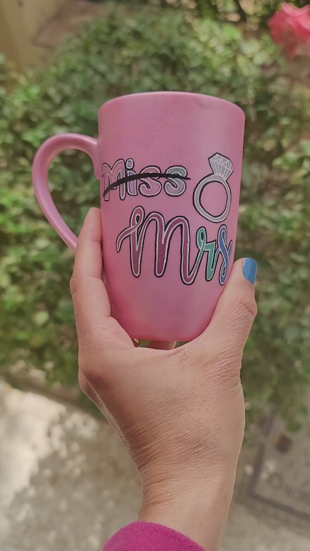 A shimmery pink hand-painted mug with the word miss crossed out and the words mrs in larger font, with a diamond ring drawn on the side. and on the other side the name "youmna" and a little crown on the letter Y