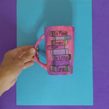 A Pink hand painted mug with books on top of each other and words written in between making the sentence