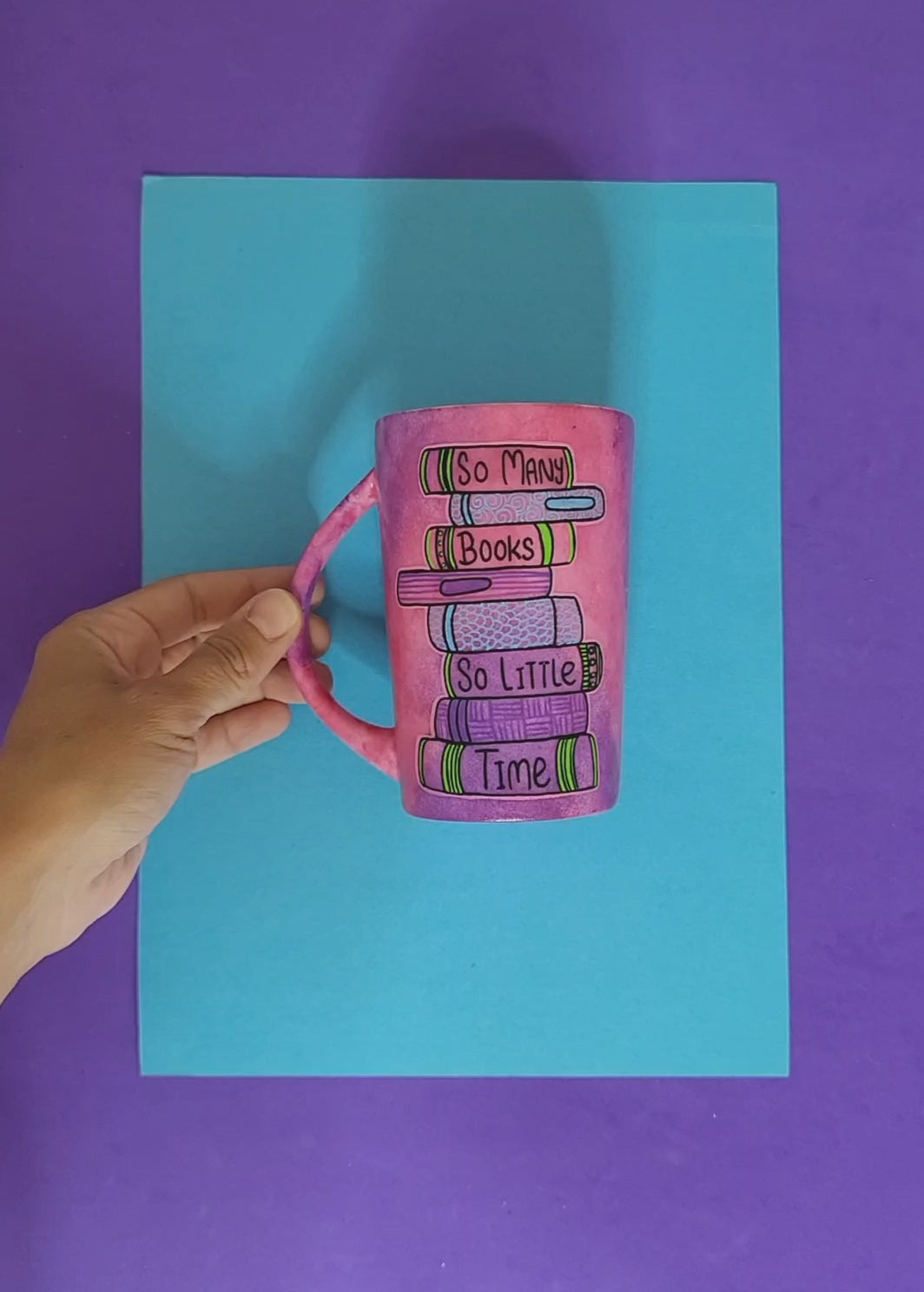 A Pink hand painted mug with books on top of each other and words written in between making the sentence" So Many books, So little time" and the other side has the name "Dee"