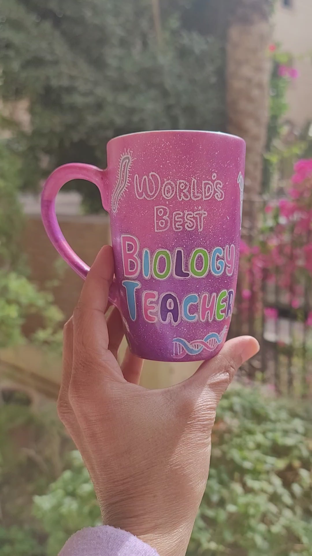 A Pink Galaxy hand painted mug with the words " worlds best biology teacher" and biology items scattered all over (DNA, Plant cell, microscope...etc.) and the other side has the name "Mariam"