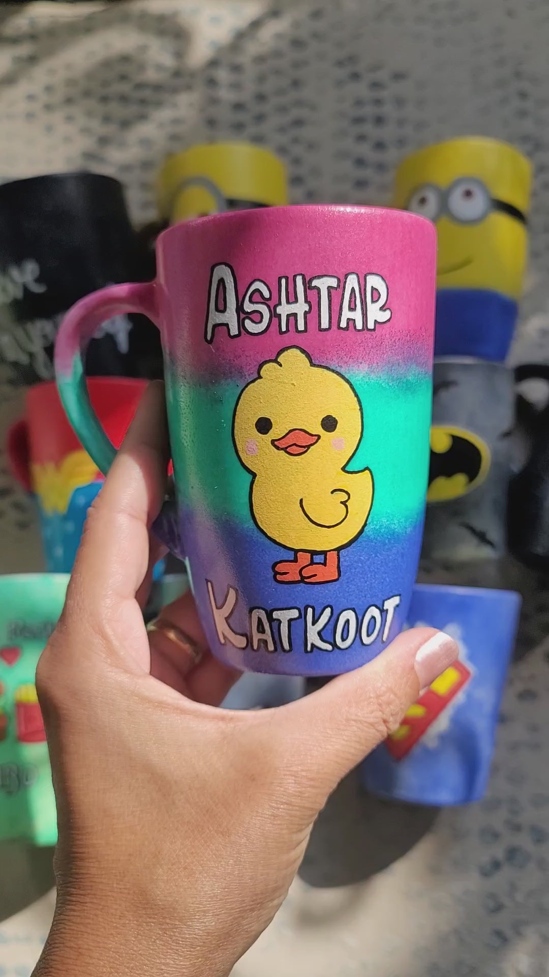 A gradient Hand apinted mug (pink- green and blue) with the word "Ashtar" on top the the duck painted in the center than the word "Katkoot" below it and the other side has the name "nada"