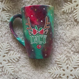 A Christmas galaxy hand painted mug with reindeer antlers with a red bow and reindeer ears, then below it the name 