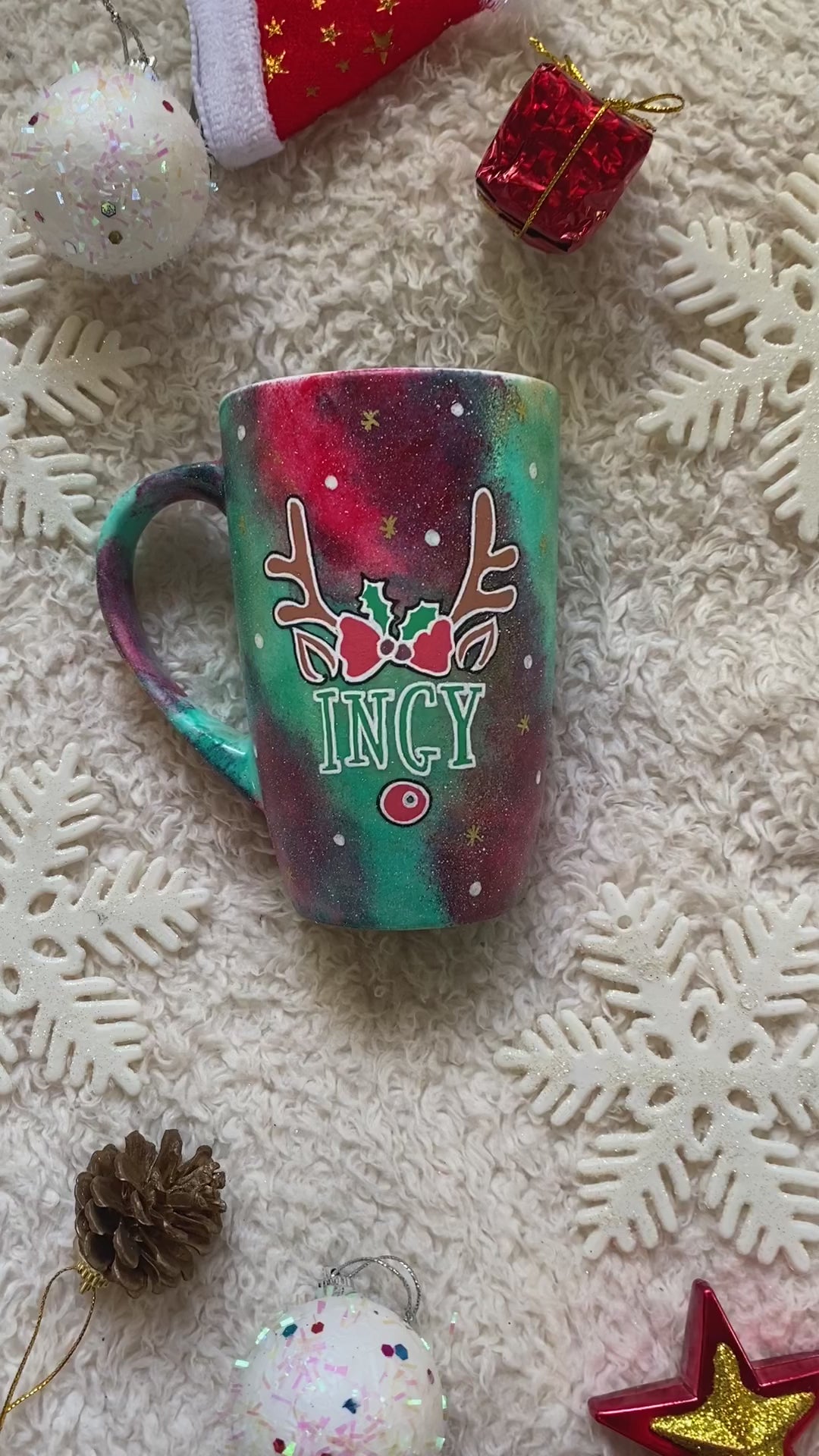 A Christmas galaxy hand painted mug with reindeer antlers with a red bow and reindeer ears, then below it the name "Ingy" and below it the reindeer nose, with white dots and gold stars scattered all over the mug. and the other side a tree drawn in white and colored in green with a gold star on top