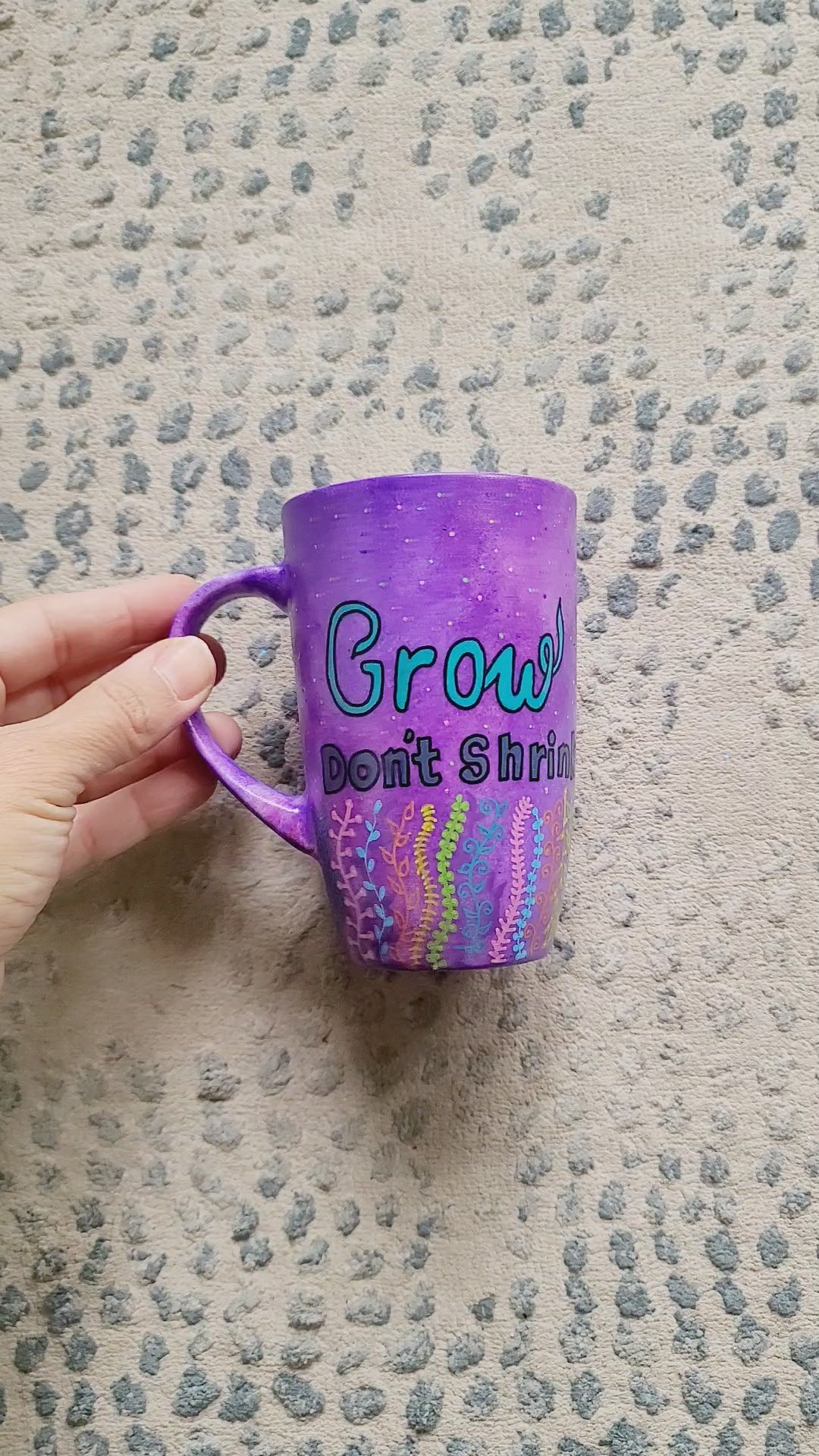 A Purple hand painted mug with the words "grow, Don't Shrink" and plants rising from the bottom of the mug to the third of height all around