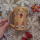 A Gold hand painted mug with gnomes all around the bottom half of the mug and festive lighting all over the top of the mug and the name nadodo in the center and other side has a stocking hung from the lights with the letter N