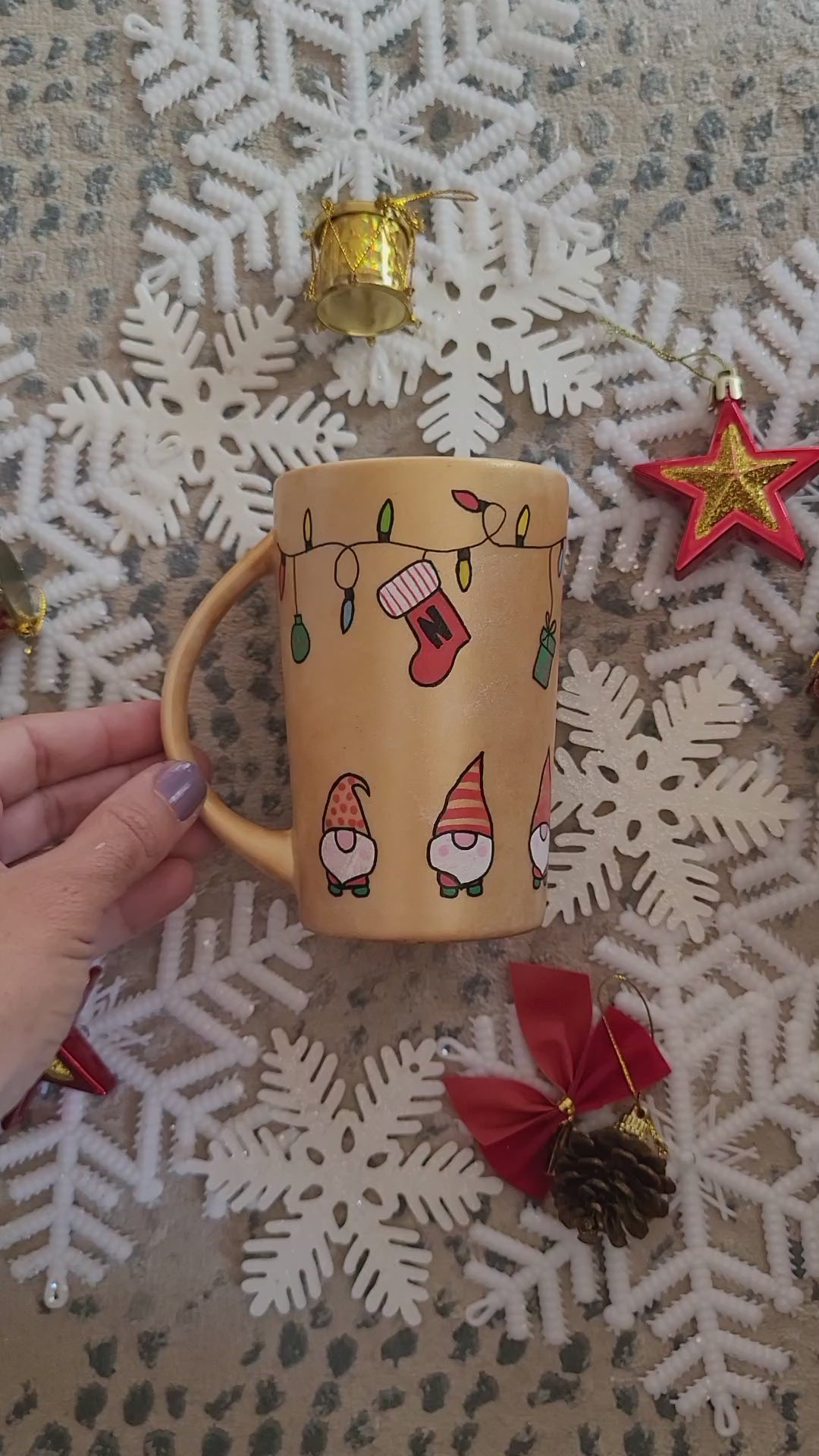 A Gold hand painted mug with gnomes all around the bottom half of the mug and festive lighting all over the top of the mug and the name nadodo in the center and other side has a stocking hung from the lights with the letter N