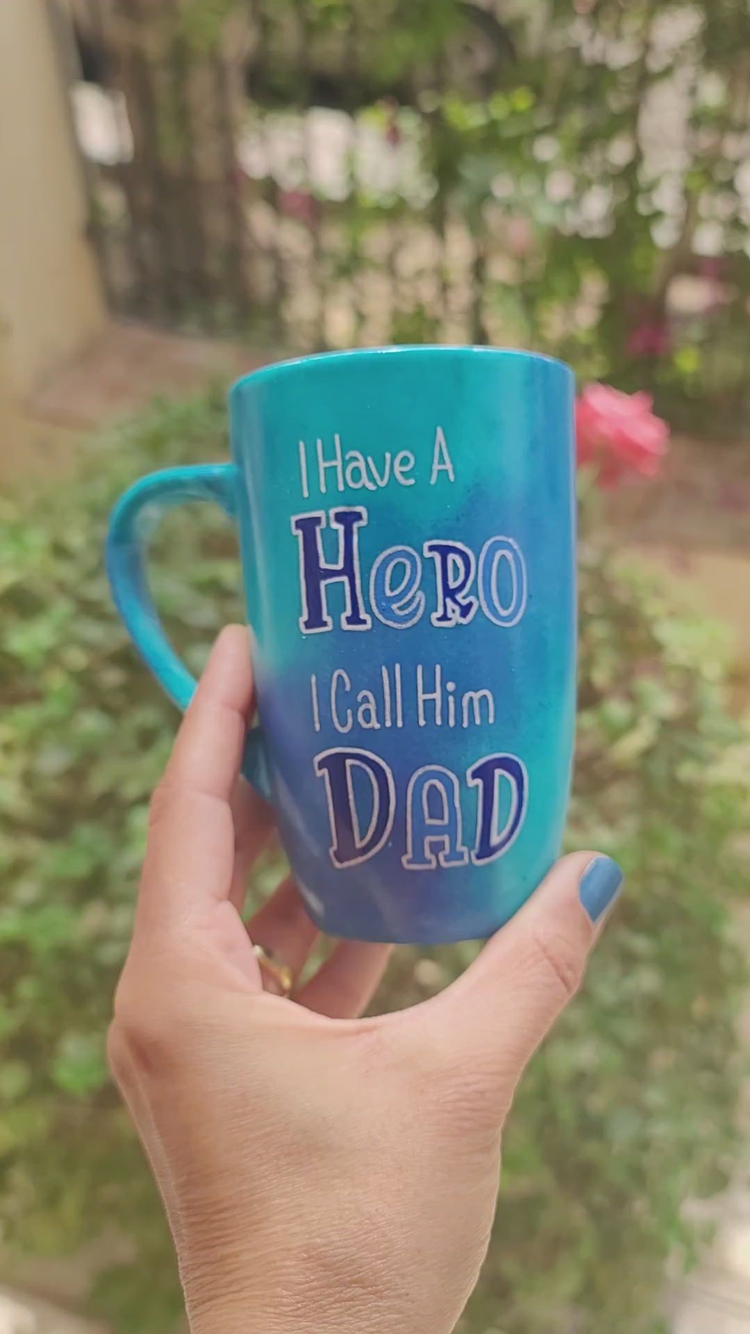 A blue hand-painted mug with the words "I Have a Hero. I Call Him Dad" and the words Hero and Dad are in larger font and colored and the other side has the name "Ahmed"