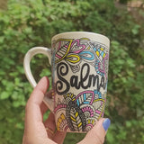 A Pearl white hand painted mug with dooldes drawn all over in black and colored in pinks, yellow and blue, with the name 