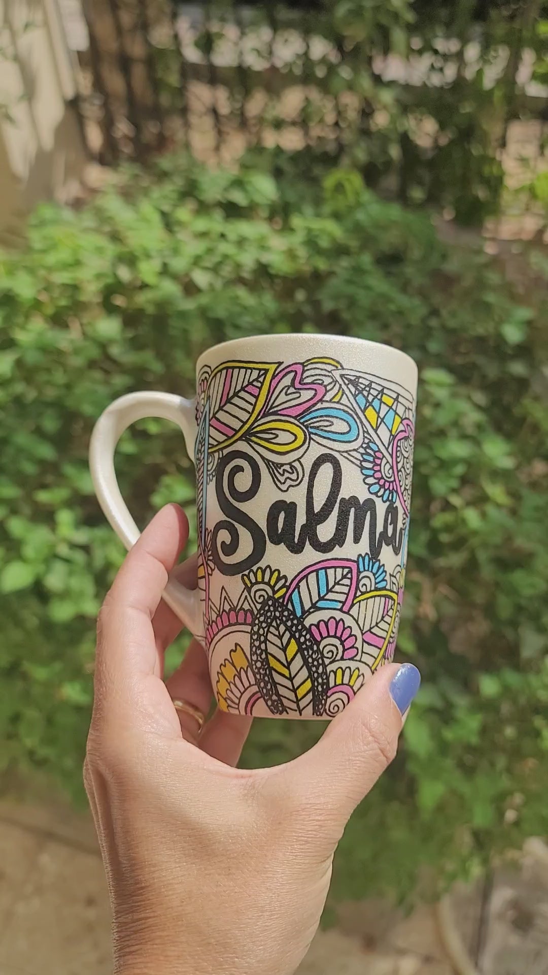 A Pearl white hand painted mug with dooldes drawn all over in black and colored in pinks, yellow and blue, with the name "Salma" written in between