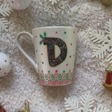 A Pearl white mug with the letter D written filled with doodles in black and colored in green and red. then hz patterns drawn all through the bottom of the mug. with green and red dots, and gold stars scattered all over. and the sentence 