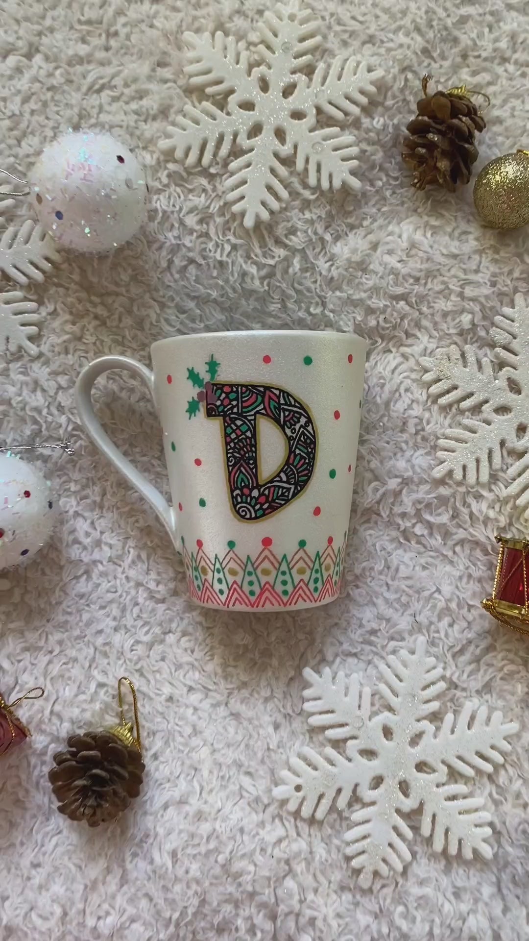 A Pearl white mug with the letter D written filled with doodles in black and colored in green and red. then hz patterns drawn all through the bottom of the mug. with green and red dots, and gold stars scattered all over. and the sentence "Diaa's Christmas mug" written on the other side