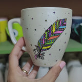 A Pearl white hand painted mug with a colorful feather outlined in black and filled with paterns in colors of orange, yellow, pink, purple, green and blue and the name 